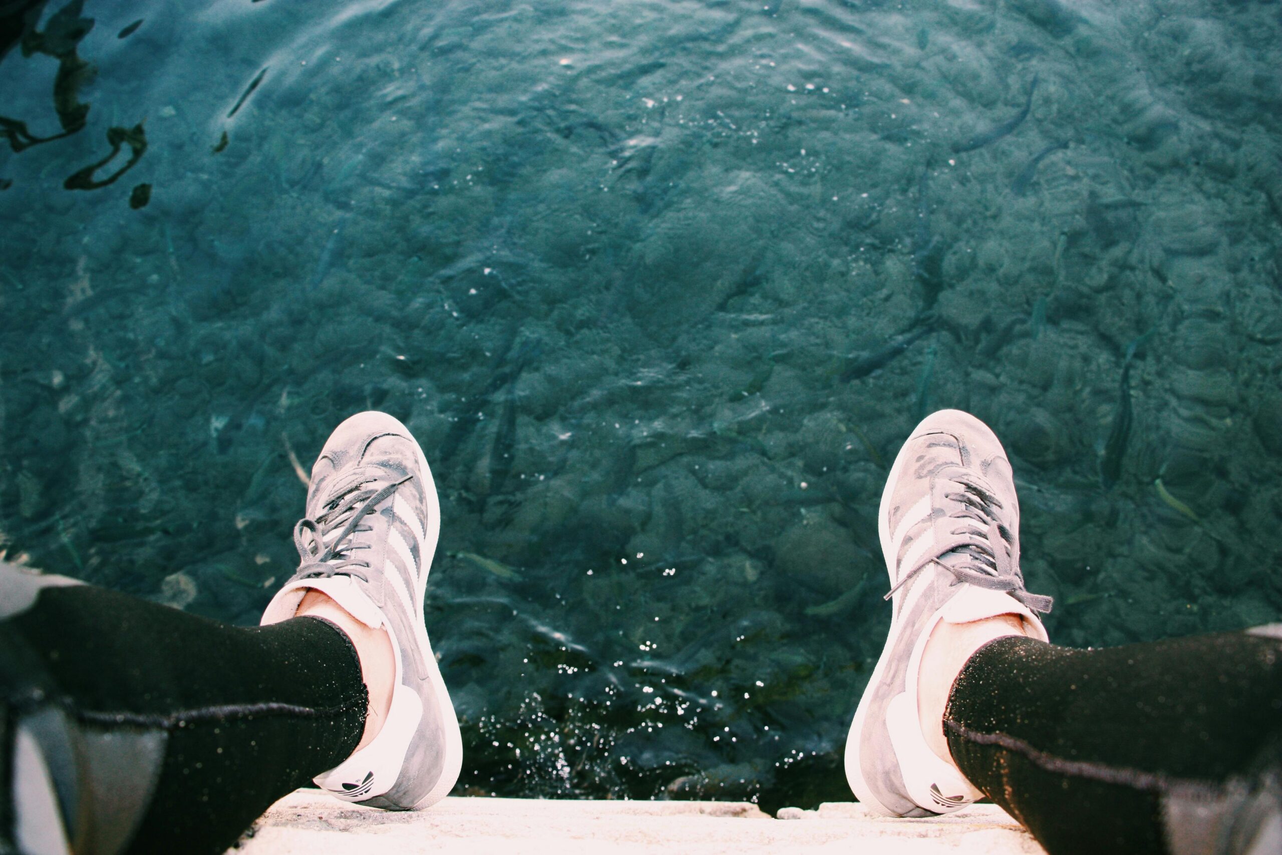 Feet in sneakers dangle over a clear blue waterbody, capturing an outdoor leisure moment.