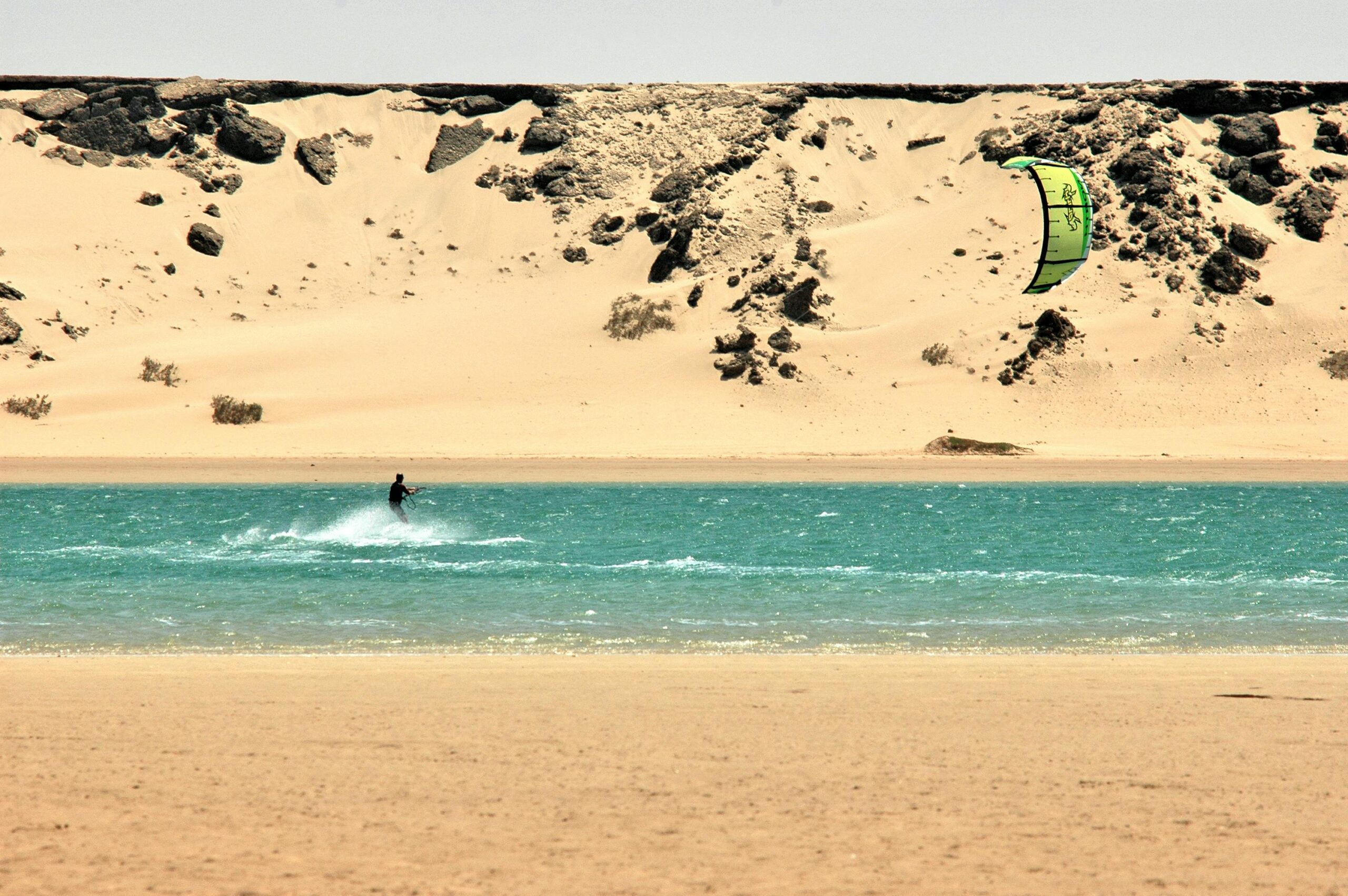 Kitesurfer enjoys waves against stunning Dakhla dune backdrop.